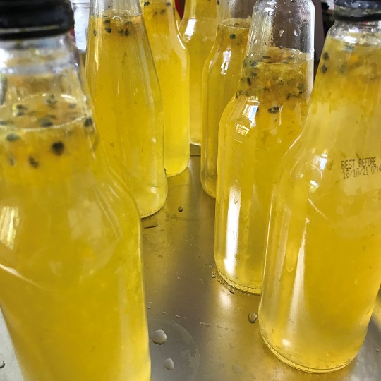 A number of small re-purposed soda bottles without labes filled with bright yellow liquid cordial, small flecks of passion fruit seeds can be seen at top of bottles, bottles have black lids